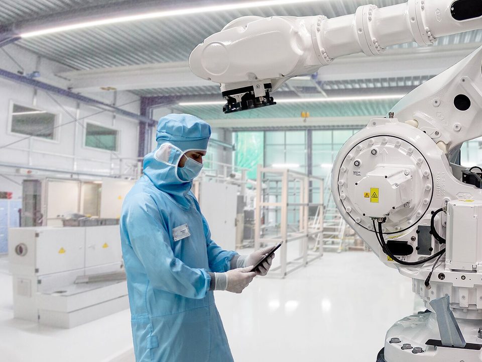 Engineer in a cleanroom suit checking a tablet beside a robot arm in a bright cleanroom